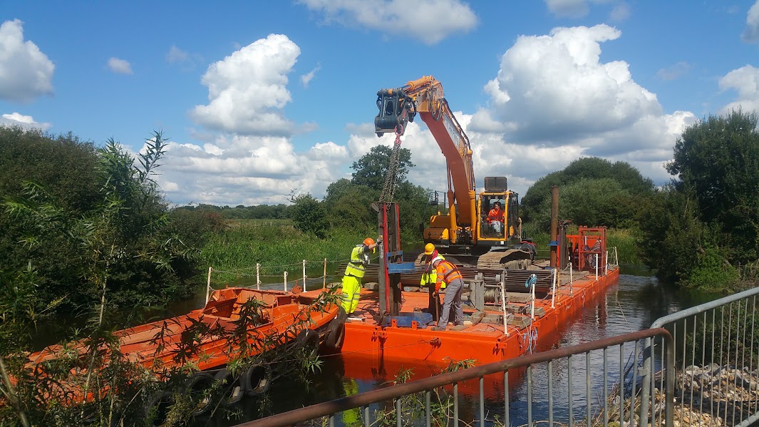 Excavator and workers installing sheet piles from a floating platform on a waterway