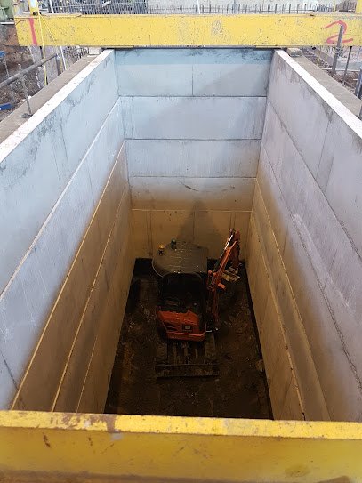 Small excavator digging within a supported rectangular shaft lined with concrete panels
